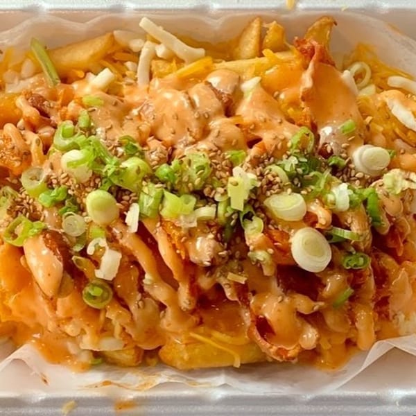 Close-up of loaded fries with orange sauce, cheese, green onions, and sesame seeds in a white container.