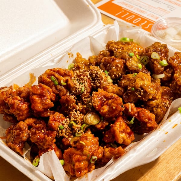 A takeout container filled with Korean fried chicken, garnished with sesame seeds and green onions.