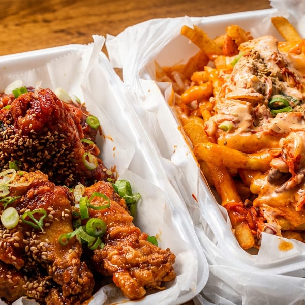 Two takeout containers with Korean fried chicken wings and kimchi fries topped with sauce and green onions.