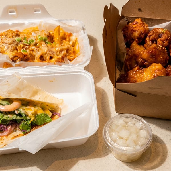 Takeout containers filled with cheesy fries, a taco, and Korean fried chicken, with a side of pickled radish.