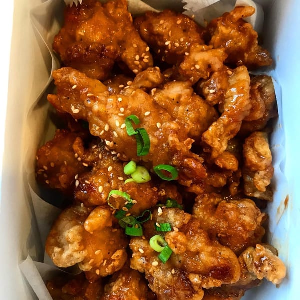 Close-up of crispy orange chicken pieces, garnished with sesame seeds and chopped green onions in a white box.