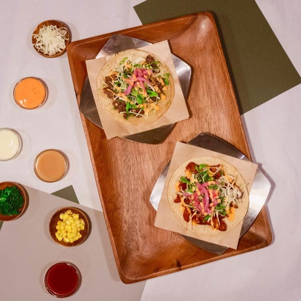 Two tacos with various toppings and small bowls of sauces and ingredients on a wooden tray.