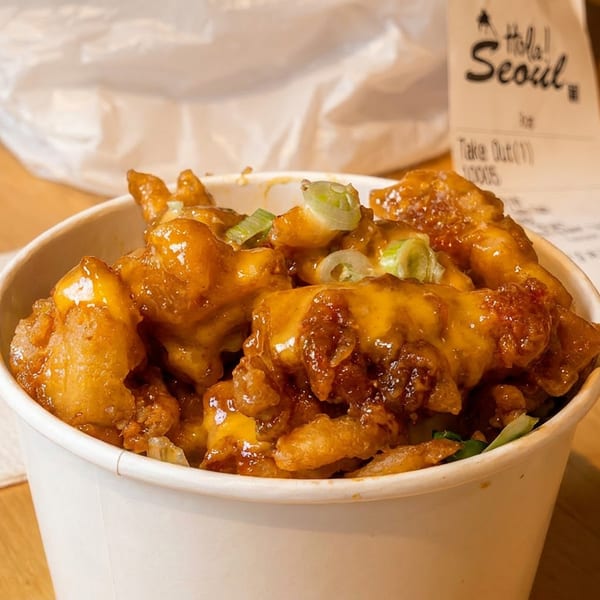 A white paper bowl filled with crispy Korean fried chicken topped with melted cheese and green onions.