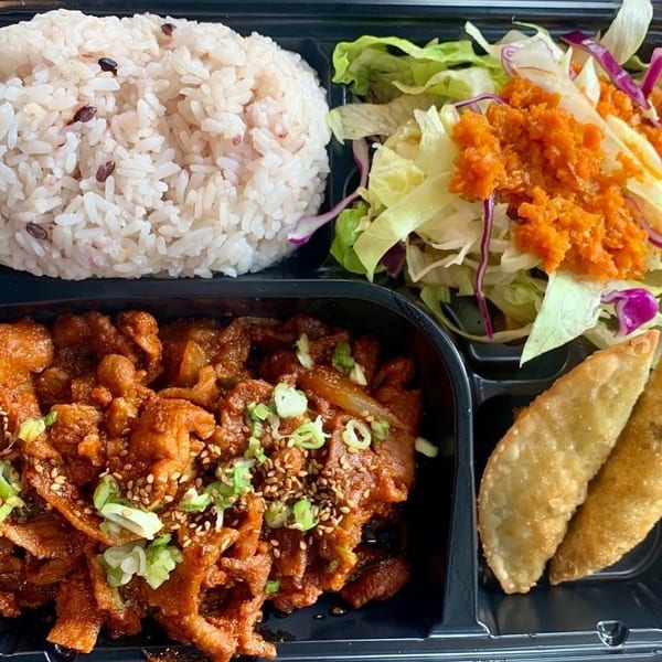 A bento box with rice, spicy pork, salad with orange dressing, and two fried dumplings.