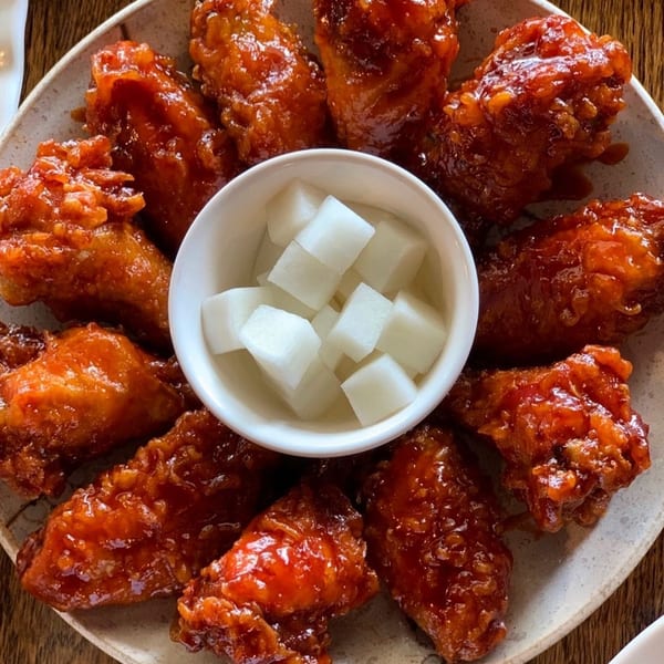 A plate of glossy, red-orange Korean fried chicken wings surrounds a small bowl of white pickled radish cubes.
