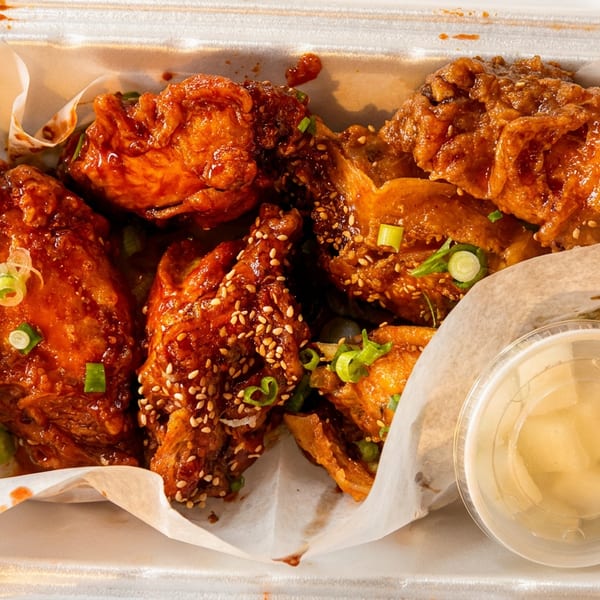 Sweet and spicy Korean fried chicken wings with sesame seeds and scallions in a takeout container.